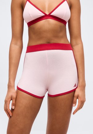 Torso féminin portant un short de sport rose clair et un brassard assorti avec une bordure rouge, debout les bras détendus le long du corps.
