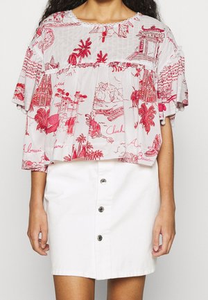 Kobieta ubrana w biały krótki top z czerwonymi tropikalnymi i architektonicznymi nadrukami oraz białą mini spódnicę z guziczkami na przodzie.