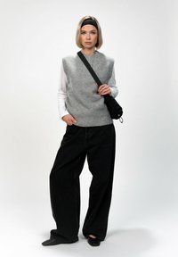 Maglione senza maniche grigio in lana sopra una maglietta bianca a maniche lunghe, abbinato a jeans neri a gamba larga e scarpe flat nere, mentre porta una borsa nera.