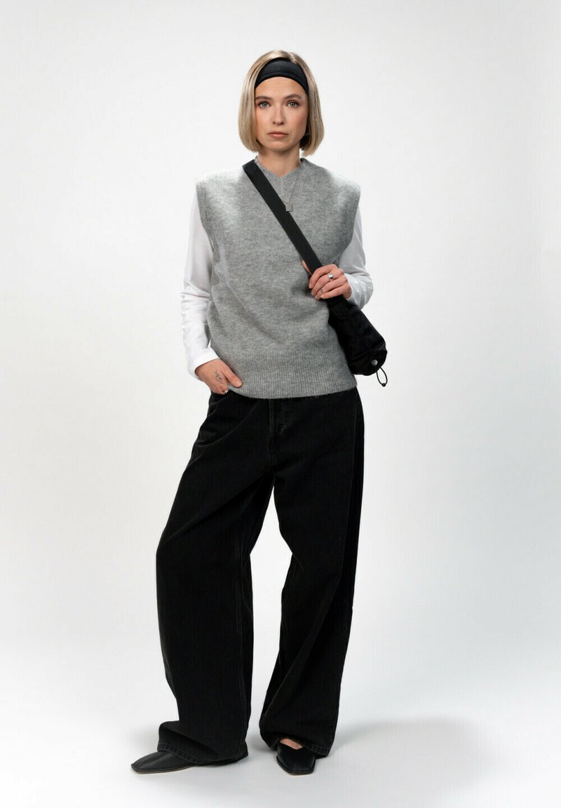 Maglione senza maniche grigio in lana sopra una maglietta bianca a maniche lunghe, abbinato a jeans neri a gamba larga e scarpe flat nere, mentre porta una borsa nera.