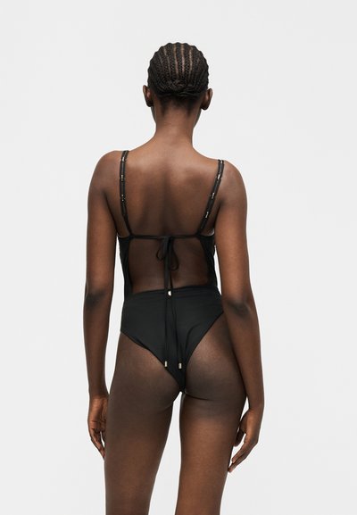 Γυναίκα με πλεκτό μαλλί, φοράει μαύρο ολόσωμο μαγιό με διάφανο πίσω μέρος και δεμένες τιράντες, στέκεται μπροστά σε απλό φόντο.