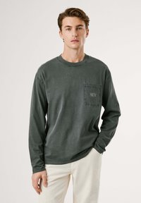 Camiseta de algodón gris de manga larga con cuello redondo, que presenta un bolsillo en el pecho y detalles de costura. Llevada con pantalones de color claro.