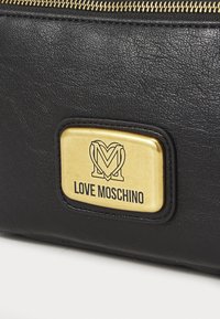 Borsa in pelle nera con una placca rettangolare dorata recante il testo "LOVE MOSCHINO" e le iniziali decorative in rilievo. Dettaglio della chiusura a zip visibile.