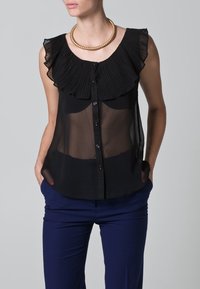 Blusa nera trasparente con colletto a rouches plissettate, chiusura con bottoni e silhouette aderente, abbinata a pantaloni blu navy. Accento con collana dorata.