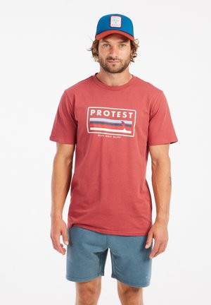 Rode t-shirt met "PROTEST" logo en blauwe strepen, gecombineerd met blauwe shorts. De pet is blauw met een rode rand en logo aan de voorkant.
