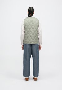 Kastēts zaļš bezmugurs, ar gludu tekstūru, uzvilkts pāri gaišai krekliņam, kombinēts ar brīviem ziliem džinsiem un brūnām kurpēm. Vienkāršs dizains, bez redzamām logo.