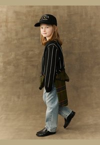 Barn som har på seg en svartstripete genser, lyseblå jeans og svarte sko, med en grønn rutete skjorte knyttet rundt livet og en svart caps.