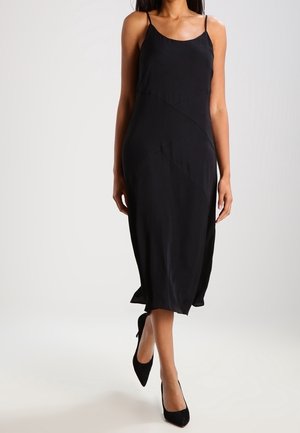 Woman wearing a sleeveless black midi dress with thin straps and black pointed high heels, walking forward against a plain background.