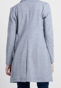 Manteau long gris en tissu texturé, coupe cintrée, avec une seule couture dans le dos. Il présente un col arrondi et des manches longues.