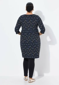 Zwarte jurk met kleurrijke verticale patronen, driekwartmouwen, elastische taille, gecombineerd met zwarte leggings en metallic platte schoenen.