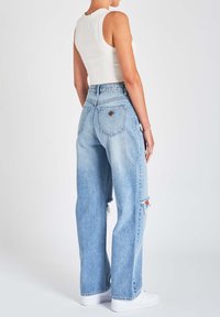 Jeans de ganga de cintura alta, azul claro, com um corte relaxado, apresentando detalhes desgastados e bolsos traseiros. Combinados com ténis brancos.