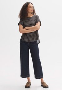 Blusa nera a maniche corte con design geometrico, abbinata a pantaloni a gamba larga in denim scuro a vita alta e sandali marroni.