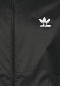 Black windbreaker jacket made of lightweight fabric, featuring a front zipper and a white embroidered Adidas logo on the left chest.