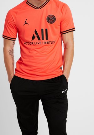 Mężczyzna w jaskrawo pomarańczowej koszulce piłkarskiej Paris Saint-Germain z czarnymi logo oraz czarnych spodniach Nike, ręce w kieszeniach.
