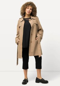 Gabardina beige con cintura ceñida, grandes botones y bolsillos laterales, llevada sobre una blusa negra y pantalones cortos negros, acompañada de zapatos negros.