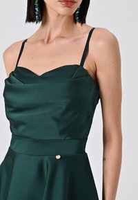 Robe en satin vert foncé avec un décolleté en cœur, des bretelles fines et une taille cintrée avec un détail doré. Texture lisse.