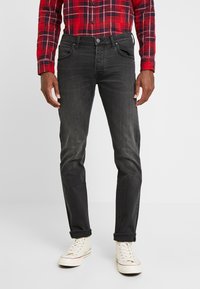 Jeans in denim nero con una vestibilità slim, cinque tasche e una finitura leggermente sbiadita; indossati con una camicia a quadri rossi e sneakers bianche.