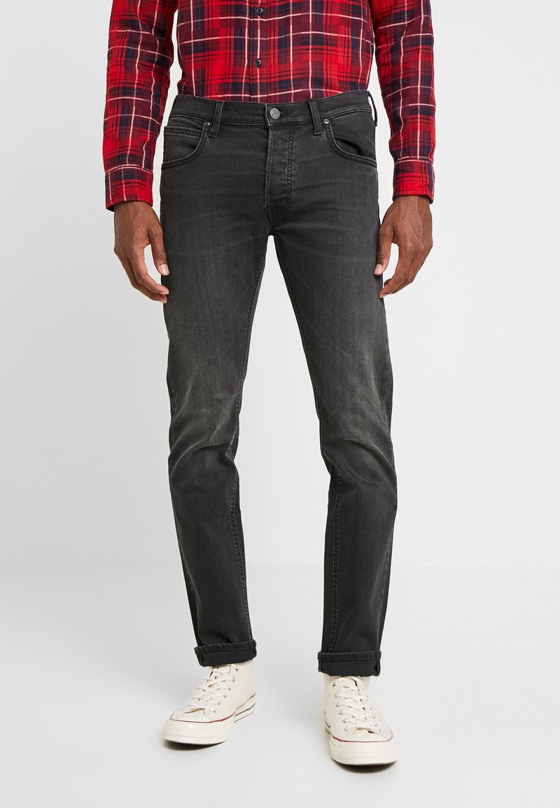 Jeans in denim nero con una vestibilità slim, cinque tasche e una finitura leggermente sbiadita; indossati con una camicia a quadri rossi e sneakers bianche.