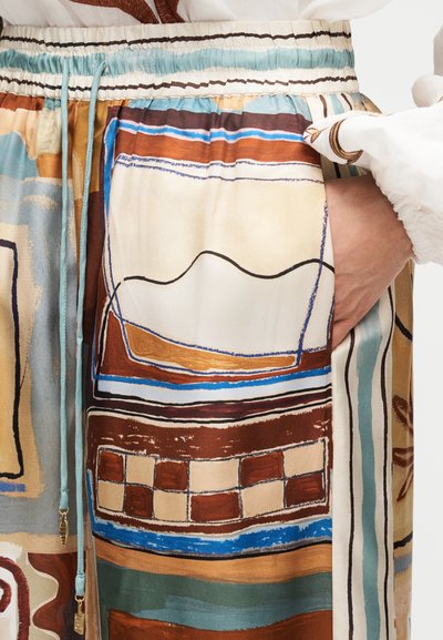 Χέρι στην τσέπη ενός φαρδιού παντελονιού με αφηρημένο γεωμετρικό μοτίβο σε γήινους τόνους με μπλε λεπτομέρειες και ελαστική μέση με κορδόνια.