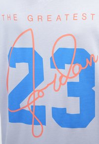 Bavlnené tričko so svetlosivým pozadím, na ktorom je výrazné modré číslo "23" a ružový text "Jordan", s nápisom "THE GREATEST" nad nimi.