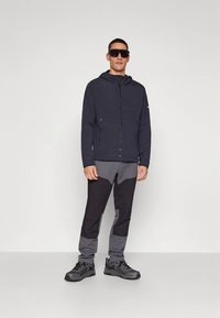 Tamsiai mėlyna striukė su užtrauktuku ir gobtuvu, derinama su pilkais ir juodais spalvų derinimo kelnėmis. Modelis dėvi saulės akinius ir lauko batus.