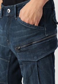 Dark denim cargo pants feature a fitted design with multiple pockets, including a zippered side pocket. The fabric has a smooth, slightly distressed texture.