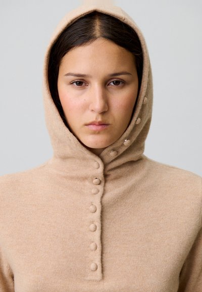 Hoodie en tricot beige avec un col montant et une patte de boutonnage à l'avant avec des boutons ronds. Texture douce et design ajusté.