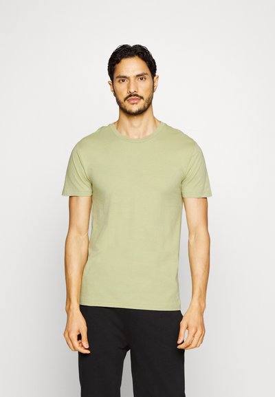 Ανοιχτό πράσινο βαμβακερό T-shirt με γύρο λαιμό και κοντά μανίκια, παρουσιασμένο σε έναν άνδρα απέναντι από έναν απλό φόντο, συνδυασμένο με μαύρο παντελόνι.