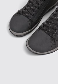 Zapatillas negras con acabado mate, punteras redondeadas, superficies texturizadas y suelas de goma gris en contraste. Presenta cordones planos negros.