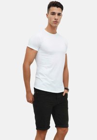 White short-sleeve t-shirt paired with black rolled hem shorts. The model has a sturdy build and is wearing a watch on his left wrist.