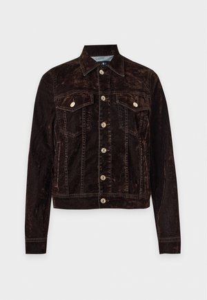 Une femme porte une veste marron foncé boutonnée sur le devant, un jean foncé et des bottes noires à talons, sur un fond clair uni.