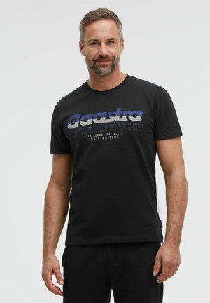 Man with short hair and beard wearing black Gaastra T-shirt with blue and white sailing tech text, standing against plain light background.