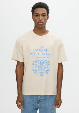 Joven con camiseta beige con diseño azul de temática mediterránea que muestra un jarrón, un pez y texto en francés, combinado con vaqueros azul claro.
