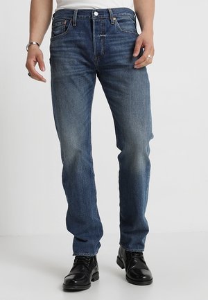 Mand iført blå straight-fit jeans og sorte læder snørestøvler, stående mod en hvid baggrund.