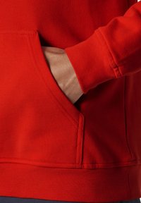 Sudadera roja con un bolsillo frontal y puños acanalados. La tela parece suave y texturizada, con costuras visibles a lo largo de las uniones.