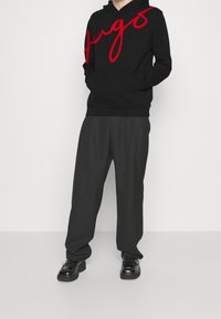 Schwarzer Kapuzenpullover mit rotem "Hugs"-Text, Kängurutasche und Zugkordelkapuze. Kombination mit grauen Hosen und schwarzen Schuhen, stehend vor einem weißen Hintergrund.