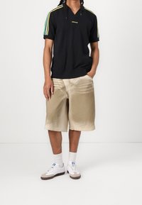 Czarna koszulka polo z krótkim rękawem z zielonymi i żółtymi paskami na ramionach, w połączeniu z jasnobeżowymi szortami o delikatnej teksturze i wzorze. Białe sneakersy.