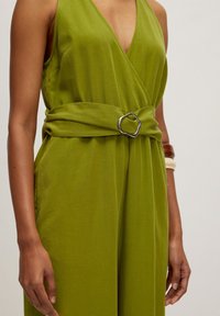 Grüner Jumpsuit mit tiefem V-Ausschnitt und einem gebundenen Gürtel, der eine silberne, geometrische Schnalle aufweist. Hergestellt aus weichem, glattem Stoff mit lockerer Passform.