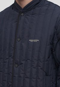 Veste matelassée bleu marine avec coutures verticales, col rond, boutons-pression à l'avant et logo "ARMANI EXCHANGE MILANO / NEW YORK" sur la poitrine.