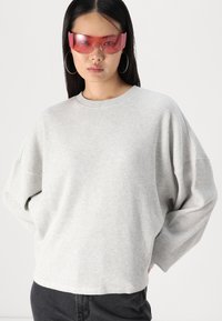 Sweater de malha cinza claro com ombros caídos, mangas largas e decote arredondado. O modelo usa óculos de sol com tonalidade rosa e grandes brincos de argola.