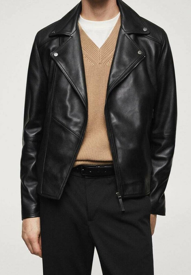Personne portant une veste en cuir noire sur un pull beige en maille à col en V et un pantalon noir avec une ceinture noire, debout, les mains détendues.