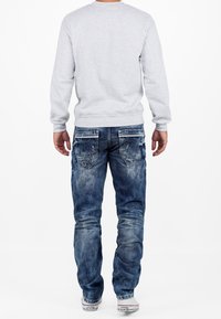 Gray sweatshirt and faded blue jeans with a distressed look, featuring back pockets and subtle branding on the right pocket. White sneakers.