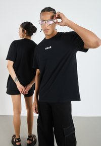 Černé oversized trička s malou bílou grafikou. Model má na sobě růžové sluneční brýle; pozadí je světlé a neutrální.