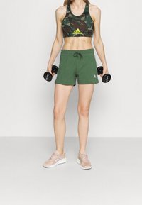 Pantalones cortos deportivos verdes combinados con un sujetador deportivo de camuflaje y zapatos beige. La modelo sostiene mancuernas negras, con una cintura ajustable y logotipos de la marca.