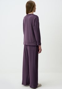 Set di abbigliamento leisure di colore viola, composto da una maglietta a maniche lunghe dal taglio rilassato e pantaloni a gamba larga, entrambi realizzati in tessuto morbido e liscio, con dettagli minimi.