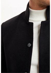 Cappotto nero in misto lana con colletto alto, dotato di chiusura a bottoni e un maglione bianco a collo alto testurizzato sotto.