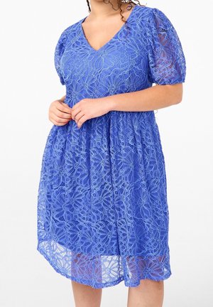 Frau trägt ein knielanges blaues Spitzenkleid mit Puffärmeln und Blumenmuster, Hände sanft in der Taille verschränkt.