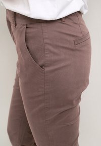 Brązowe bawełniane spodnie o gładkiej teksturze. Posiadają boczne kieszenie oraz jedną tylną kieszeń. Dopasowany krój bez widocznych zapięć ani zdobień.