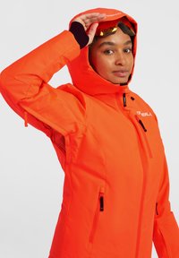 Heldere oranje waterdichte jas met een capuchon, voorzien van ritszakken en mesh ventilatieopeningen, ontworpen voor buitenactiviteiten. Gladde textuur, duurzaam materiaal.