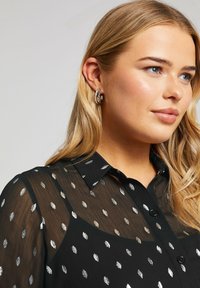 Zwarte doorzichtige blouse met zilveren bladerenpatroon, knoopsluiting aan de voorkant en kraagaccent. Model heeft los, golvend haar en ringoorbellen.